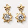A pair of gold dangle earrings featuring a star-shaped top and a sunburst lower section with a large clear round-cut gemstone, shown on a light gray background.