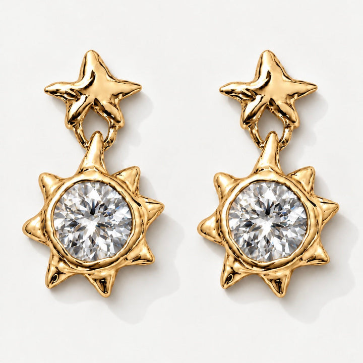 A pair of gold dangle earrings featuring a star-shaped top and a sunburst lower section with a large clear round-cut gemstone, shown on a light gray background.