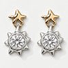 A pair of dangling earrings on a light gray background. Each earring has a gold star-shaped post with a silver-toned sunburst setting holding a clear round gemstone.