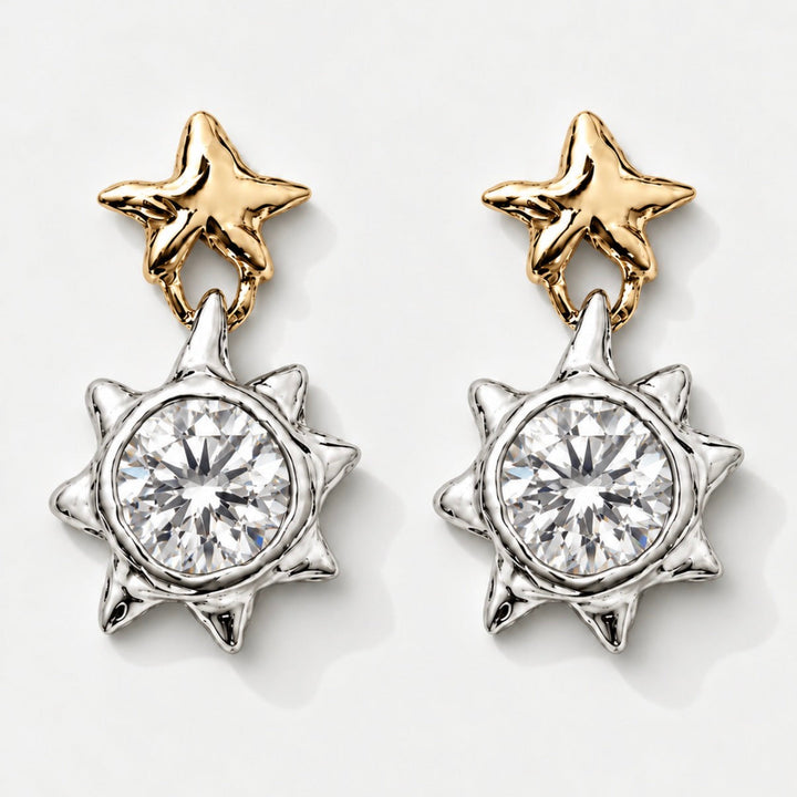 A pair of dangling earrings on a light gray background. Each earring has a gold star-shaped post with a silver-toned sunburst setting holding a clear round gemstone.