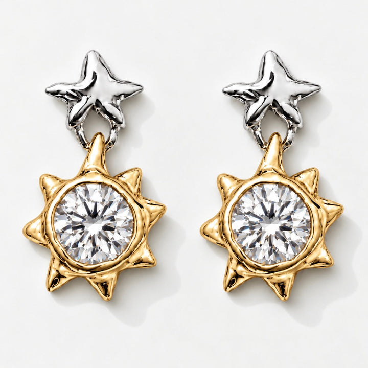 A pair of two-tone star and sun dangle earrings. Each earring features a polished silver star stud at the top and a gold-toned sunburst charm with a large clear round-cut center stone, set against a clean white background.