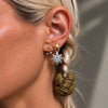 A close-up of a model's ear wearing a multi-layered celestial earring stack. The stack features a gold star stud, a silver compass rose stud, tiny clear crystal dots, and a dangling olive green fabric scrunchie.