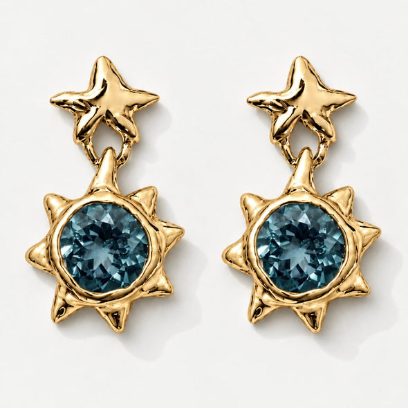 A pair of gold dangle earrings featuring a star-shaped top and a sunburst lower section with a faceted blue gemstone, shown on a clean white background.