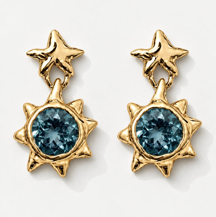 A pair of gold dangle earrings featuring a star-shaped top and a sunburst lower section with a faceted blue gemstone, shown on a clean white background.