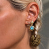 A close-up of a model's ear wearing a gold star-shaped dangle earring with a light blue gemstone and small gold studs.