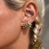 A close-up of a model's ear wearing a silver starburst stud earring with a round blue gemstone and small silver bead accents, paired with a green scrunchie in her hair.