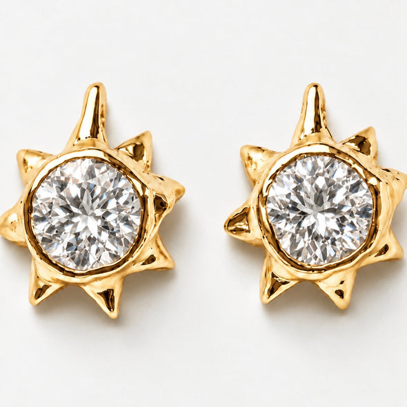 A pair of gold starburst stud earrings with a large round clear gemstone in the center, set against a plain white background.