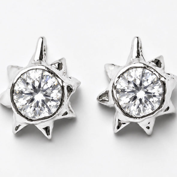 A pair of silver-toned starburst stud earrings with a clear round-cut gemstone in the center, set against a plain light gray background.