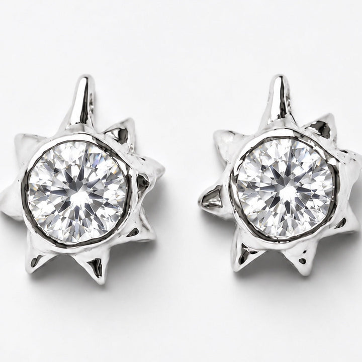 A pair of silver-toned starburst stud earrings with a clear round-cut gemstone in the center, set against a plain light gray background.