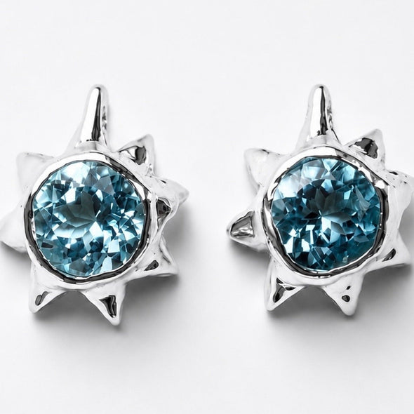 A pair of silver starburst stud earrings featuring round, faceted light blue gemstones, set against a clean white background.
