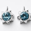 A pair of silver starburst stud earrings featuring round, faceted light blue gemstones, set against a clean white background.