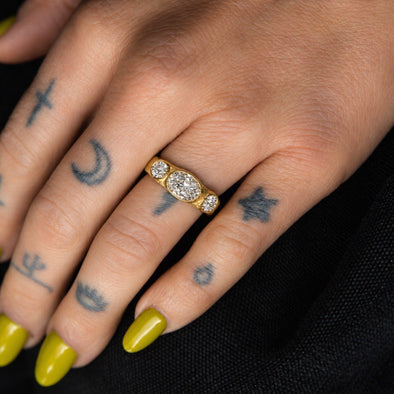 A close-up of a hand with yellow-green nail polish and black tattoos wearing a gold ring with a central round-cut diamond and smaller side diamonds, set against a dark background.