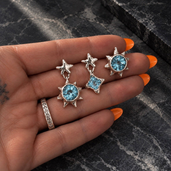 A close-up of a hand with orange nail polish holding a pair of silver star-shaped dangle earrings with round and square light blue gemstones, set against a dark gray marble background.