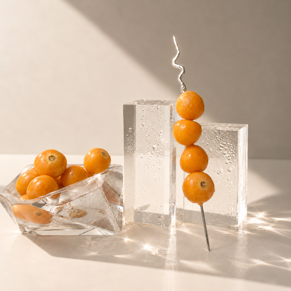 A metal skewer with four orange kumquats stands upright in a clear textured ice block, next to a glass bowl filled with kumquats on a warm beige surface with dramatic sunlight and shadows.