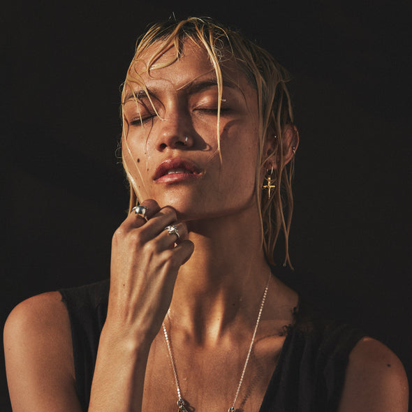 Woman with wet hair and minimal makeup wearing jewelry against a dark background