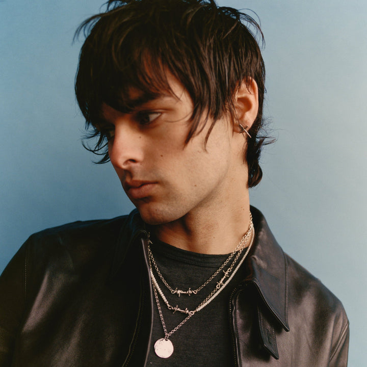 Man wearing a black leather jacket and layered necklaces against a light blue background
