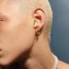 A close-up side profile of a person with short platinum blonde hair wearing a small gold hoop earring with a dangling anchor charm, paired with a pearl necklace against a dark blue-gray background.