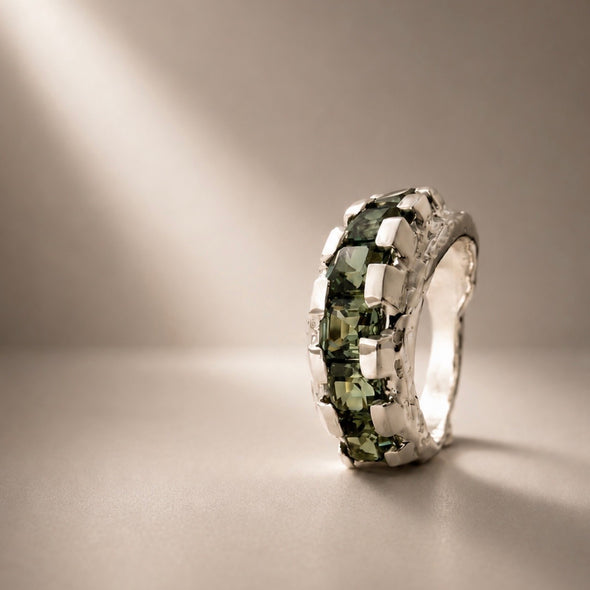 A silver ring featuring a row of green rectangular gemstones, photographed on a neutral beige surface with dramatic diagonal lighting.