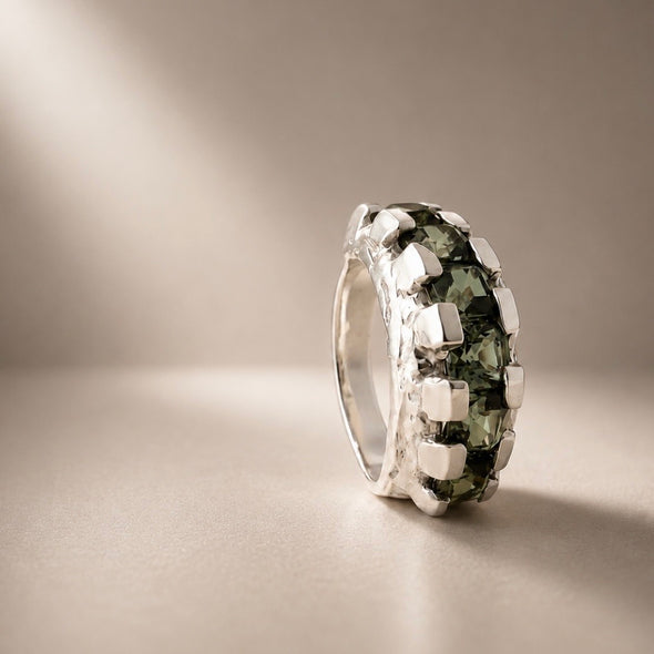 A silver ring featuring a textured, block-like setting holding dark green gemstones, photographed on a neutral beige background with soft light rays.