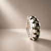 A silver ring featuring a textured, block-like setting holding dark green gemstones, photographed on a neutral beige background with soft light rays.