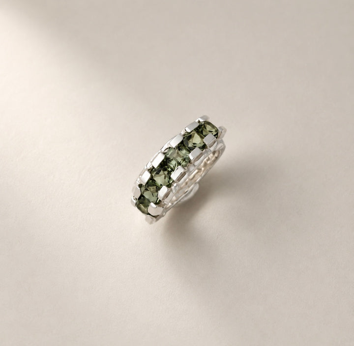 A silver band ring featuring a row of green faceted gemstones, photographed on a clean, light beige background.