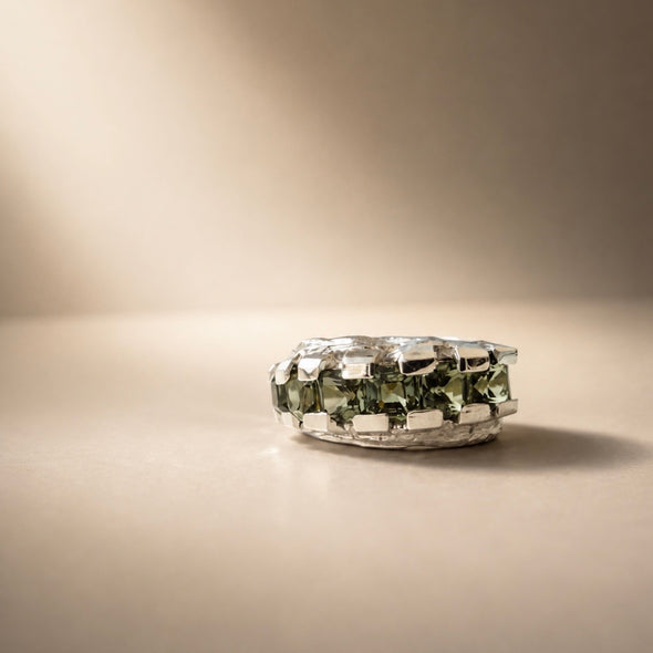 A silver ring featuring a large, faceted green gemstone, photographed on a warm beige surface with a matching background.