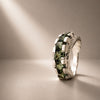 A silver ring featuring a row of green rectangular gemstones, photographed on a neutral beige surface with dramatic diagonal lighting.