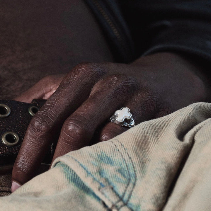 Hand wearing a ring with a cross pearl design, resting on a textured surface.