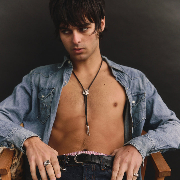 Man wearing a denim shirt and jeans sitting on a chair against a dark background