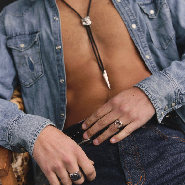 Man wearing a denim shirt with a necklace and rings, sitting against a dark background