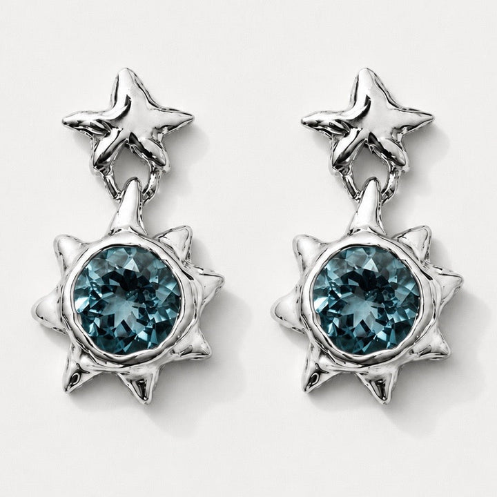 A pair of silver-toned dangle earrings featuring a small star-shaped stud at the top and a larger spiky sunburst pendant with a faceted blue-green gemstone in the center, shown on a light gray background.