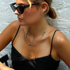 A woman with blonde-highlighted hair wearing black cat-eye sunglasses, a silver necklace, and a black spaghetti-strap top, posing against a white brick wall.