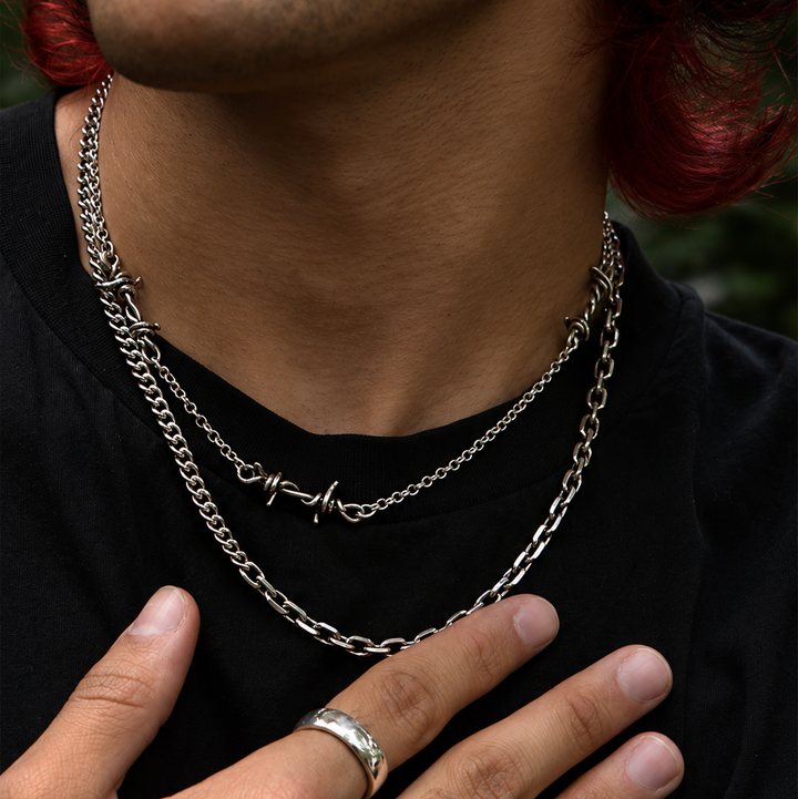 A close-up of a person wearing layered silver chain necklaces, a silver ring, and a silver-toned statement ring with a green gemstone, over a black shirt.