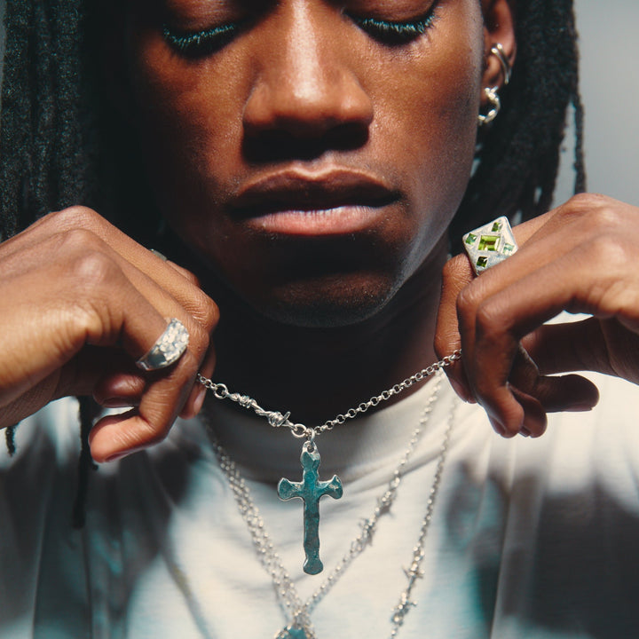 A person with a close-up on their neck, wearing a silver-colored cross-shaped pendant on a barbed chain necklace.