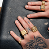 Close-up of tattooed hands wearing multiple gold rings, including chunky square, lion-head, and textured signet-style rings, resting on a dark textured leather surface.