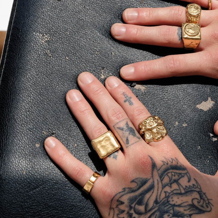 Close-up of tattooed hands wearing multiple gold rings, including chunky square, lion-head, and textured signet-style rings, resting on a dark textured leather surface.