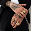 A close-up of a person's tattooed hands wearing multiple chunky gold rings, including square and textured dome designs, styled with a black blazer and a silver chain.