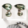 Pair of green gemstone earrings on a white card with a shell and pearl necklace below.