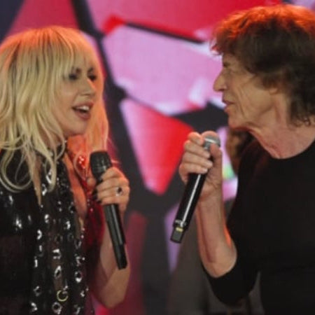 Two people holding microphones on stage, with a colorful abstract background and one person wearing a black sequined jacket and the other wearing a black top.