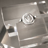 A silver ring featuring a circular, textured seal-like face with an embossed symbol, displayed on a clear acrylic stand.