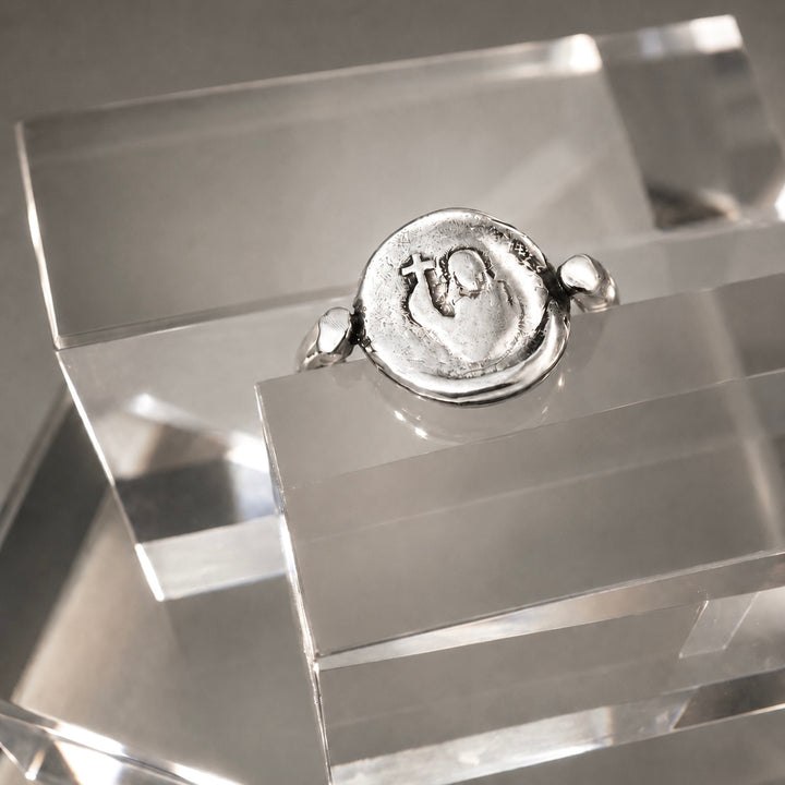 A silver ring featuring a circular, textured seal-like face with an embossed symbol, displayed on a clear acrylic stand.