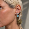 Close-up of a woman's ear wearing a silver-toned star and crescent moon earring with a light blue gemstone, alongside other small stud earrings.