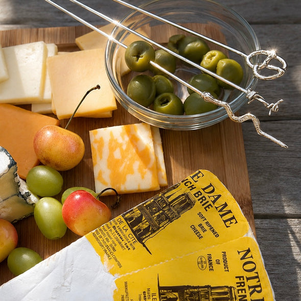 A styled cheese board scene on a wooden surface with sliced cheeses, green olives in a glass bowl, grapes, and two cheese knives with decorative handles. A yellow paper tag reading 'Notre Dame French' is visible in the foreground.
