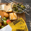 A styled cheese board scene on a wooden surface with sliced cheeses, green olives in a glass bowl, grapes, and two cheese knives with decorative handles. A yellow paper tag reading 'Notre Dame French' is visible in the foreground.