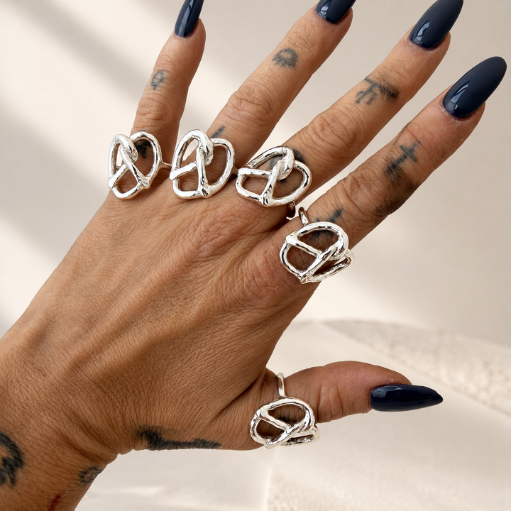 A hand with dark blue manicured nails and tattoos wearing five silver Pretzel rings, each featuring a twisted pretzel knot design, set against a warm, softly lit cream background.